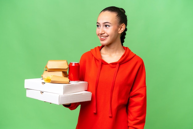 Young Arab woman holding fast food over isolated background looking to the side and smiling