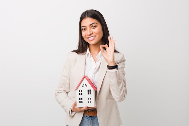 Young arab woman holding a house icon cheerful and confident showing ok gesture.