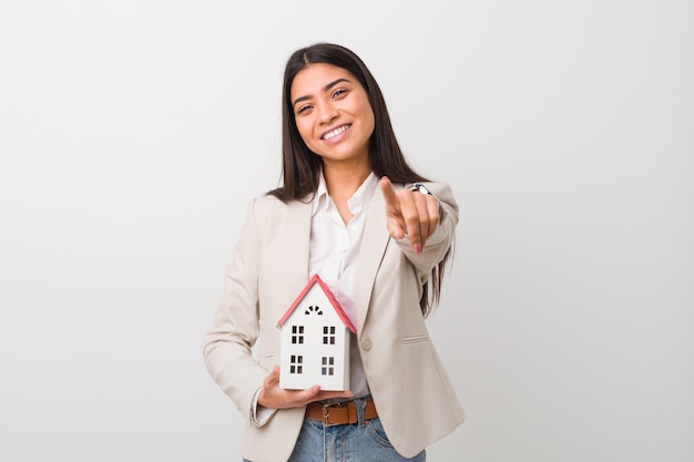 Young arab woman holding a house icon cheerful smiles pointing to front.