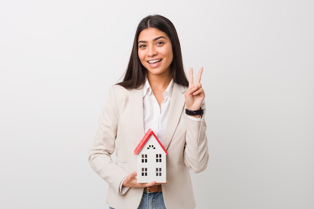 Young arab woman holding a house icon showing victory sign and smiling broadly.