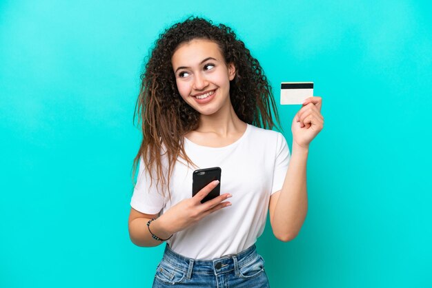 Young Arab woman isolated on blue background buying with the mobile with a credit card while thinking