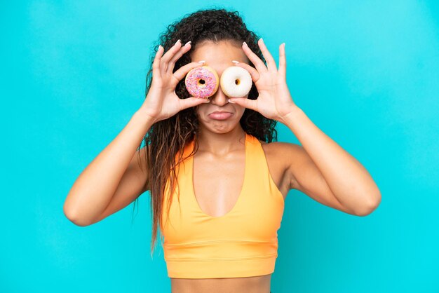 Young Arab woman isolated on blue background holding donuts in eyes with sad expression