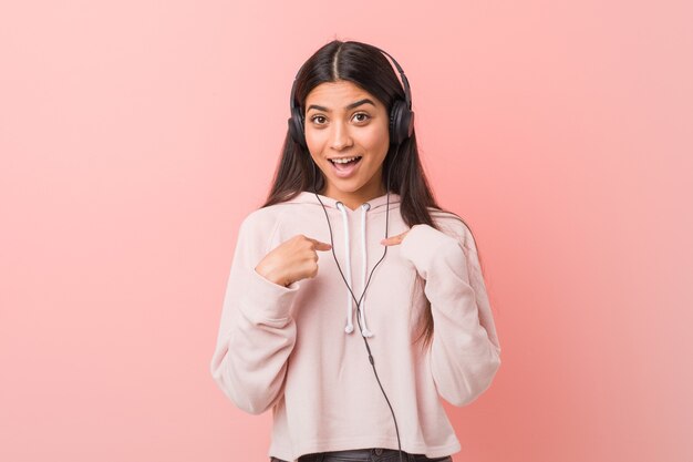 Young arab woman listening to music surprised pointing at himself, smiling broadly.