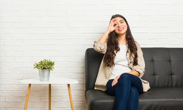 Young arab woman sitting on the sofa excited keeping ok gesture on eye.