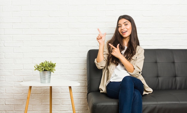 Young arab woman sitting on the sofa pointing with forefingers to a copy space, expressing excitement and desire.