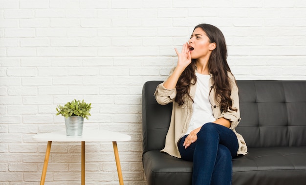 Young arab woman sitting on the sofa shouting and holding palm near open mouth.