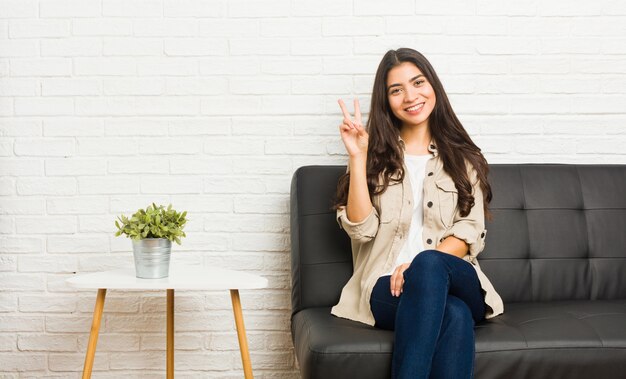 Young arab woman sitting on the sofa showing victory sign and smiling broadly.