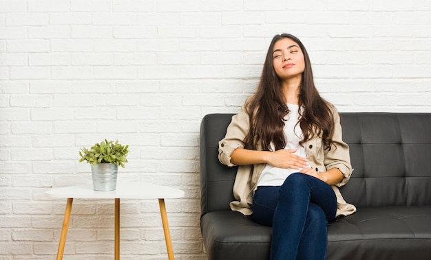 Young arab woman sitting on the sofa touches tummy, smiles gently, eating and satisfaction concept.