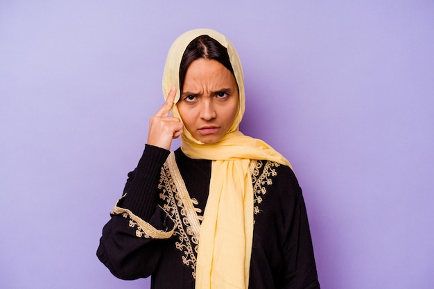 Молодая арабская женщина в типичном арабском костюме изолирована на фиолетовом фоне, указывая пальцем на храм, думая, сосредоточенная на задаче.