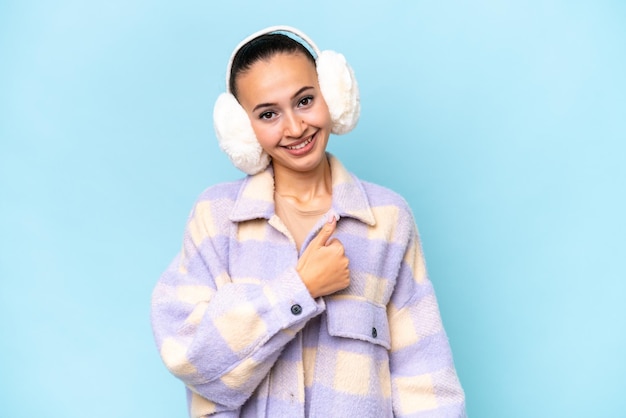 Young arab woman wearing winter muffs isolated on blue background giving a thumbs up gesture