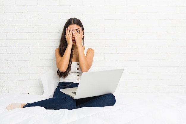 Young arab woman working with her laptop on the bed, blinks through fingers, frightened and nervous