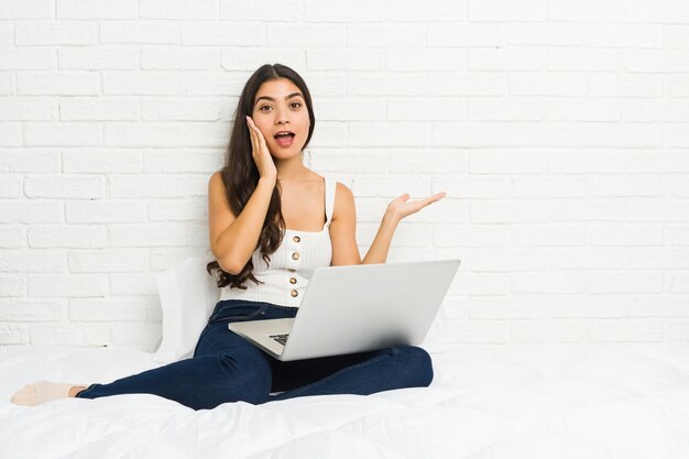 Young arab woman working with her laptop on the bed holds copy space on a palm, keep hand over cheek. Amazed and delighted.