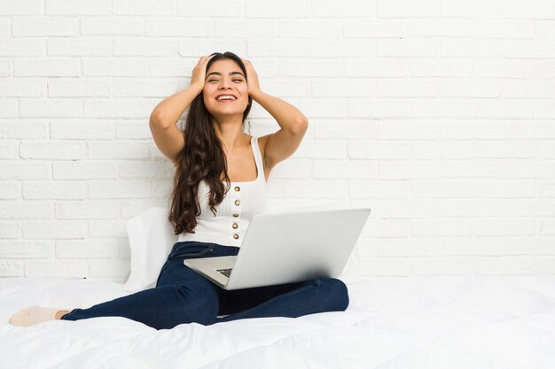 Young arab woman working with her laptop on the bed laughs joyfully keeping hands on head. Happiness concept.