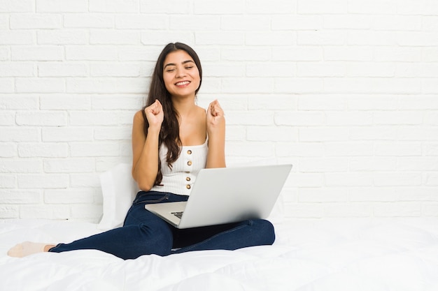 Young arab woman working with her laptop on the bed raising fist, feeling happy and successful. Victory concept.