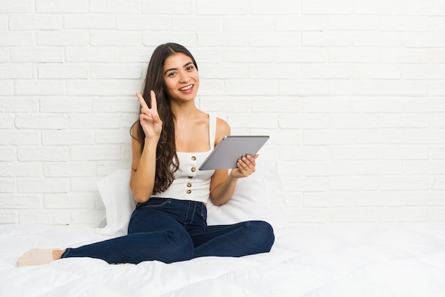 Giovane donna araba che lavora con il suo computer portatile sul letto che mostra il segno di vittoria e che sorride ampiamente.