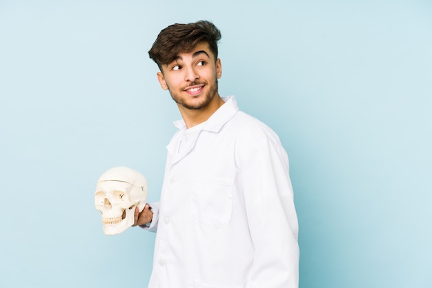 Young arabian doctor man holding a skull looks aside smiling, cheerful and pleasant.