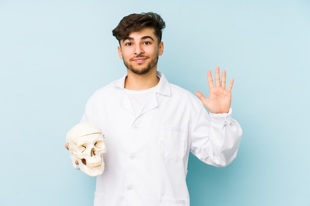 Молодой арабский доктор мужчина держит череп, улыбаясь веселый, показывая номер пять пальцами.
