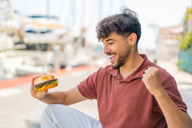 Young Arabian handsome man holding a burger at outdoors celebrating a victory