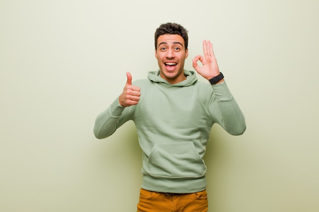Young arabian man feeling happy, amazed, satisfied and surprised, showing okay and thumbs up gestures, smiling against flat wall