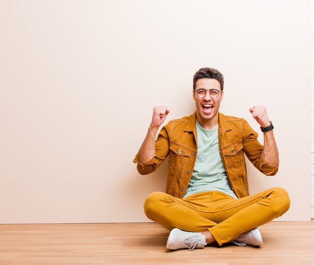 Young arabian man feeling happy, positive and successful, celebrating victory, achievements or good luck sitting on the floor