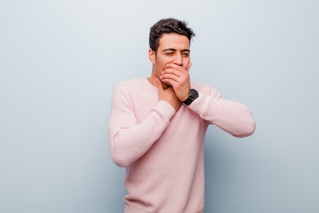 Young arabian man feeling ill with a sore throat and flu symptoms, coughing with mouth covered against gray wall