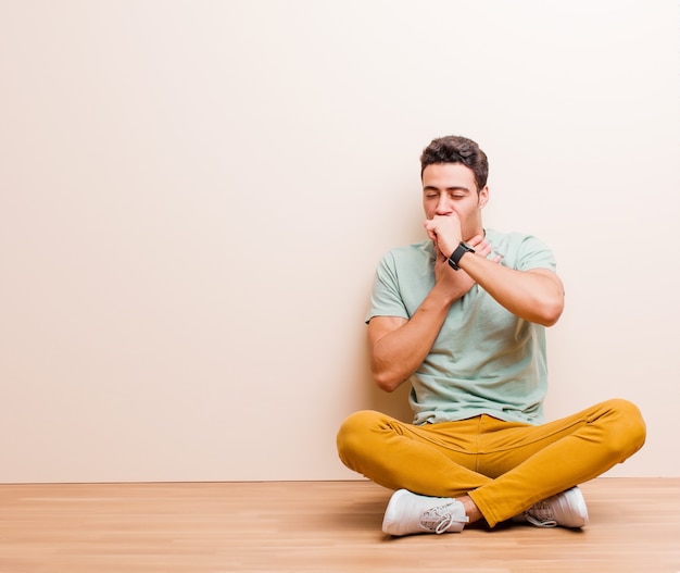 Giovane uomo arabo sentirsi male con mal di gola e sintomi influenzali, tossendo con la bocca coperta seduta sul pavimento