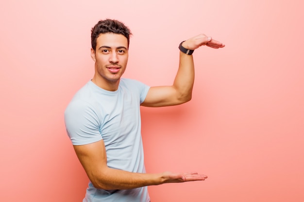 Young arabian man holding an object with both hands on side copy space, showing, offering or advertising an object against pink wall