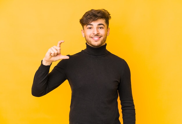 Young arabian man isolated on a yellow wall holding something little with forefingers, smiling and confident.