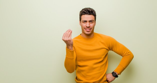 Young arabian man making capice or money gesture, telling you to pay your debts! against flat wall
