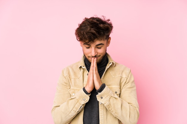 Young arabian man posing in a wall isolated holding hands in pray near mouth, feels confident