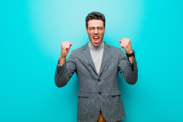 Young arabian man shouting aggressively with an angry expression or with fists clenched celebrating success against blue wall