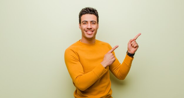 Young arabian man smiling happily and pointing to side and upwards with both hands showing object in copy space against flat wall