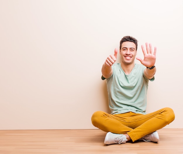 Young arabian man smiling and looking friendly, showing number six or sixth with hand forward, counting down sitting on the floor