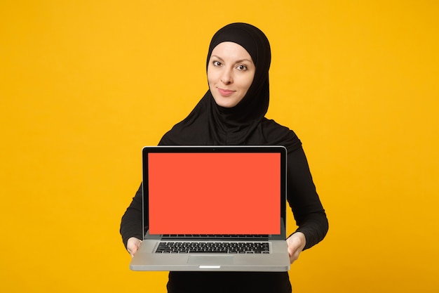 Young arabian muslim employee woman in hijab black clothes hold and work laptop pc computer isolated on yellow wall  portrait. People religious lifestyle concept. 