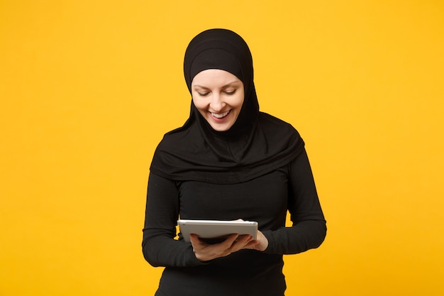 La giovane donna musulmana araba dell'impiegato in vestiti neri di hijab tiene e lavora il computer del pc della compressa isolato sul ritratto giallo della parete. concetto di stile di vita religioso della gente.