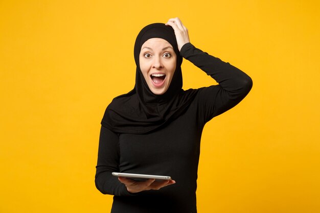 La giovane donna musulmana araba dell'impiegato in vestiti neri di hijab tiene e lavora il computer del pc della compressa isolato sul ritratto giallo della parete. concetto di stile di vita religioso della gente.