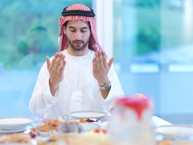 young arabian muslim man making traditional prayer to God, keeps hands in praying gesture before iftar dinner with family representing modern islam fashion and ramadan kareem concept