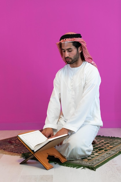 Молодой арабский мусульманин в традиционной одежде читает священную книгу Коран на молитвенном ковре перед ужином ифтар во время праздника рамадан дома.