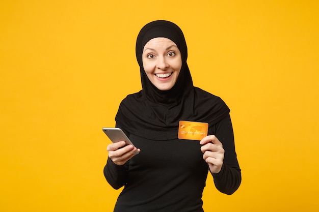 Young arabian muslim woman in hijab black clothes hold in hand mobile phone, credit card isolated on yellow wall  portrait. People religious lifestyle concept. 