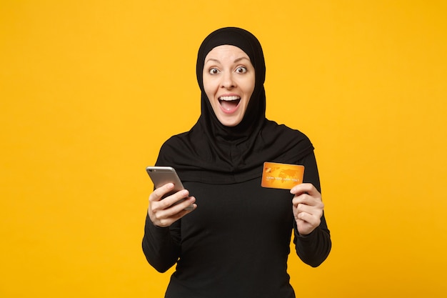 Young arabian muslim woman in hijab black clothes hold in hand mobile phone, credit card isolated on yellow wall portrait. people religious lifestyle concept