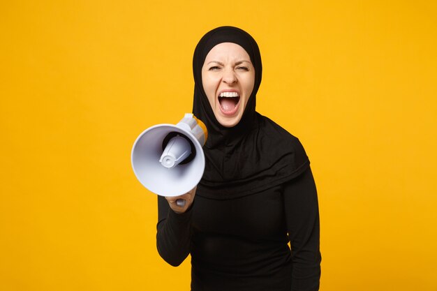 Young arabian muslim woman in hijab black clothes holds in hand bullhorn public address megaphone isolated on yellow wall portrait. People religious lifestyle concept. 