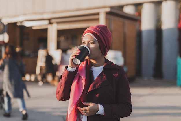 Young Arabic beautiful woman in hijab walking at street tapping on smartphone and drinking coffee