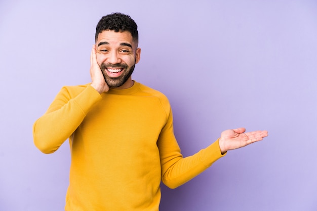 Young arabic man holds copy space on a palm, keep hand over cheek. Amazed and delighted.