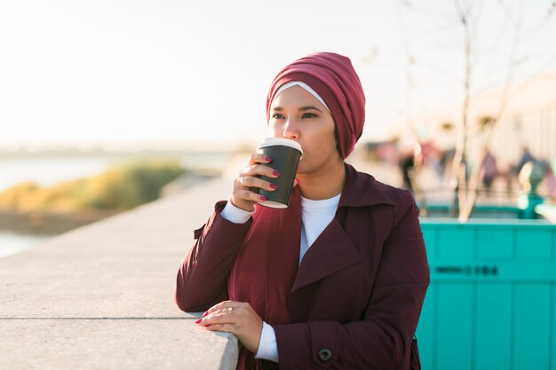 Young Arabic muslim woman in hijab standing at street seafront and drinking coffee take away copy space and place for advertising