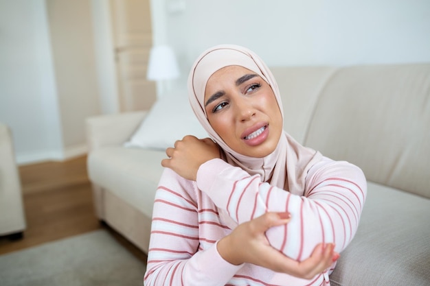 ヒジャブを被った若いアラビア人イスラム教徒女性が自宅のソファに座り、肘の痛みに苦しみながら肘に触れている