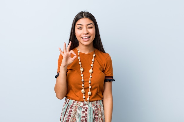 Young arabic woman winks an eye and holds an okay gesture with hand.