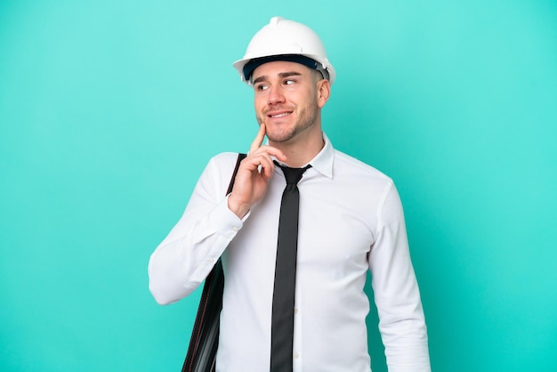 Young architect caucasian man with helmet and holding\
blueprints isolated on blue background thinking an idea while\
looking up