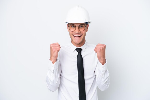 Young architect caucasian man with helmet and holding blueprints isolated on white background celebrating a victory in winner position