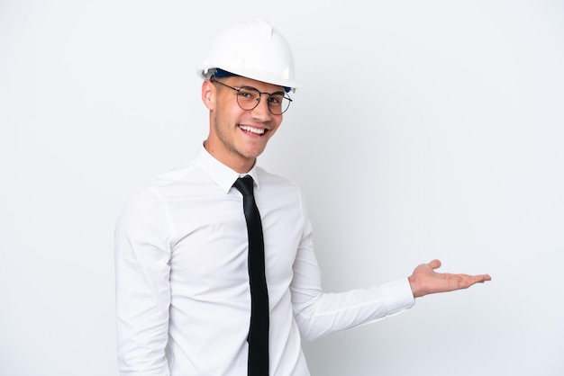 Young architect caucasian man with helmet and holding\
blueprints isolated on white background extending hands to the side\
for inviting to come