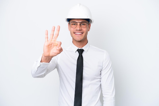 Молодой архитектор кавказец в шлеме и с чертежами на белом фоне счастлив и считает три пальцами